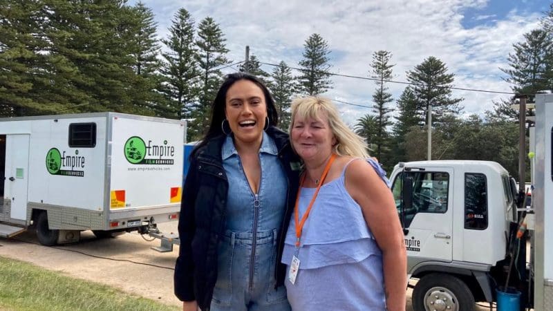 Two women smiling beside lorries with tall pine trees in the background, filming a 1 Day Home Away Tour by Celebtime Tours for fans.