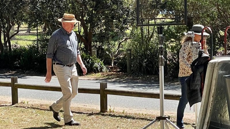 A man in a hat strolls on green grass during a 1 Day Home Away Tour, with outdoor kit and lush trees visible in the background.