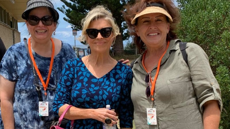 Three happy women on a Celebtime Tours 1 Day Home Away Tour, posing outside amid lush greenery and a scenic building backdrop.