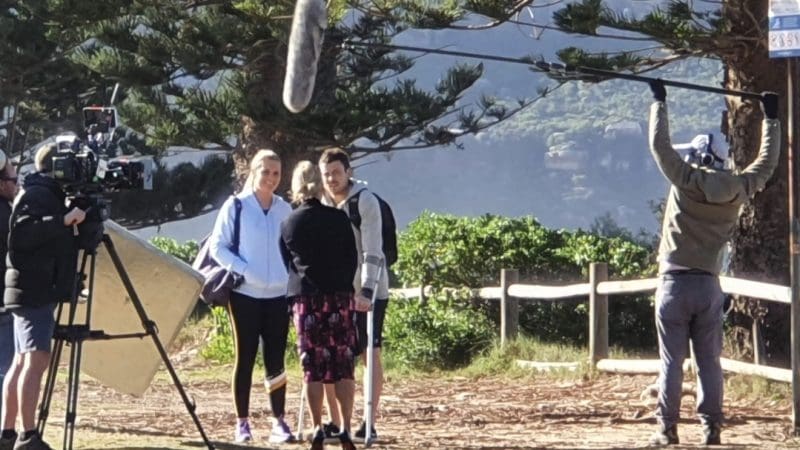 A professional film crew captures a 1 Day Home Away Tour with two participants outdoors, surrounded by lush trees and a wooden fence.