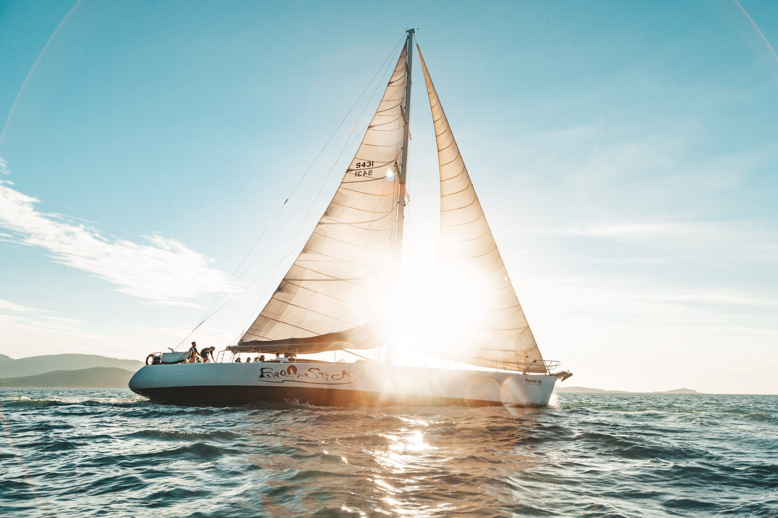 Experience the Broomstick 2 Day 2 Night Whitsunday Sailing boat gliding at sunset, golden sunlight streaming through sleek white sails.