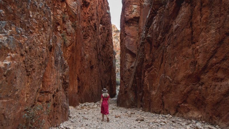 Experience the breathtaking 4 Day Uluru to West MacDonnell Tour as a woman in a red dress explores vibrant red canyon cliffs.