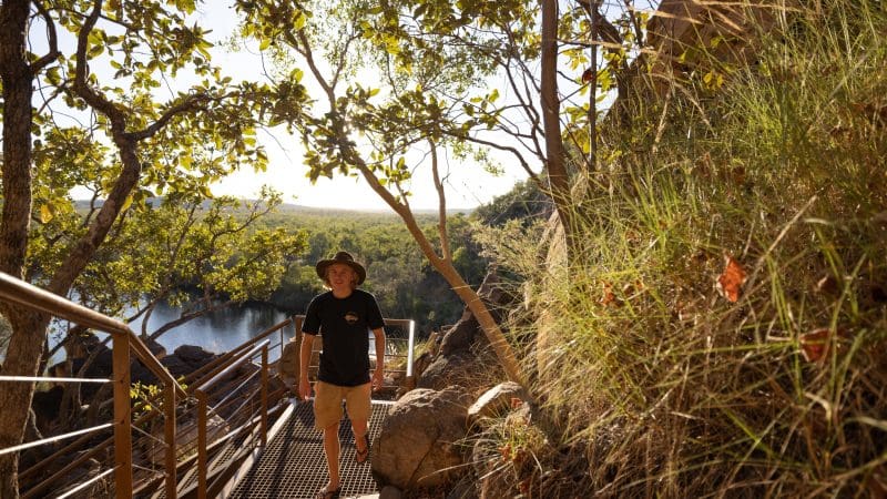Traveller in a sun hat ascends a metal staircase surrounded by lush trees and rocky terrain beside Katherine Gorge river on 1 Day Cruise.