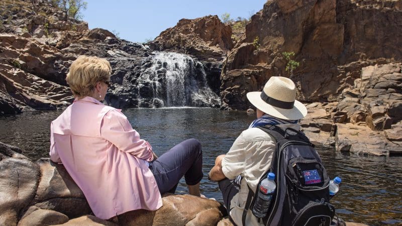 Two people relaxing by Edith Falls admire stunning waterfalls and rugged cliffs on a Katherine Gorge Cruise day trip from Darwin.