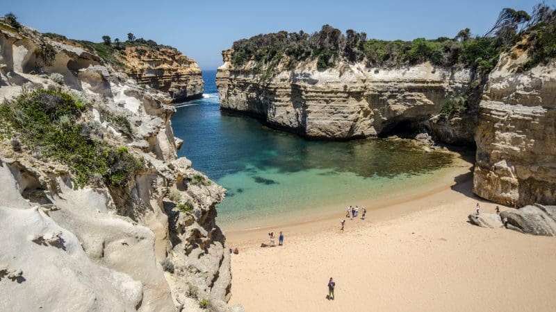 Stunning sandy beach with vibrant turquoise water, dramatic cliffs, and clear blue sky on Great Ocean Road Sunset Tour from Melbourne.