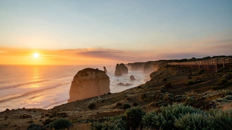 Stunning sunset illuminates rugged ocean rocks and a scenic promenade on a 1 Day Great Ocean Road Sunset Tour from Melbourne, Australia.