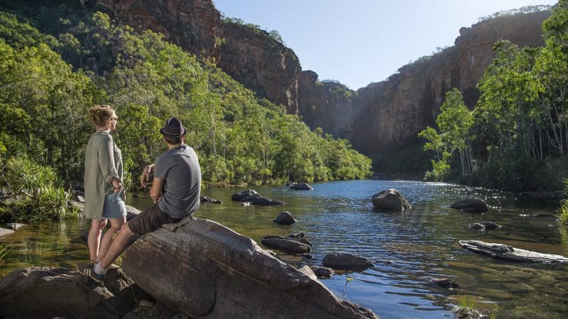 Explore the scenic 5 Day Kakadu, Katherine Gorge & Litchfield 4WD Adventure from Darwin as two people relax riverside on sunlit rocks.