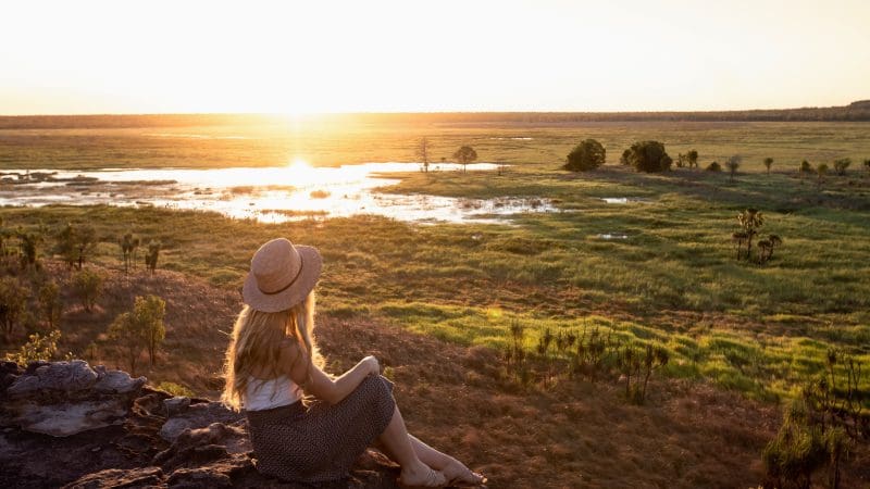 Woman wearing a hat sits on a rock, watching sunset on a 5 Day Kakadu, Katherine Gorge & Litchfield 4WD tour from Darwin.