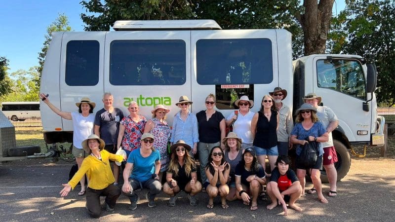 Happy tourists pose in front of a white tour coach before their 3 Day Kakadu and Litchfield National Park 4WD Safari Tour on a sunny day.