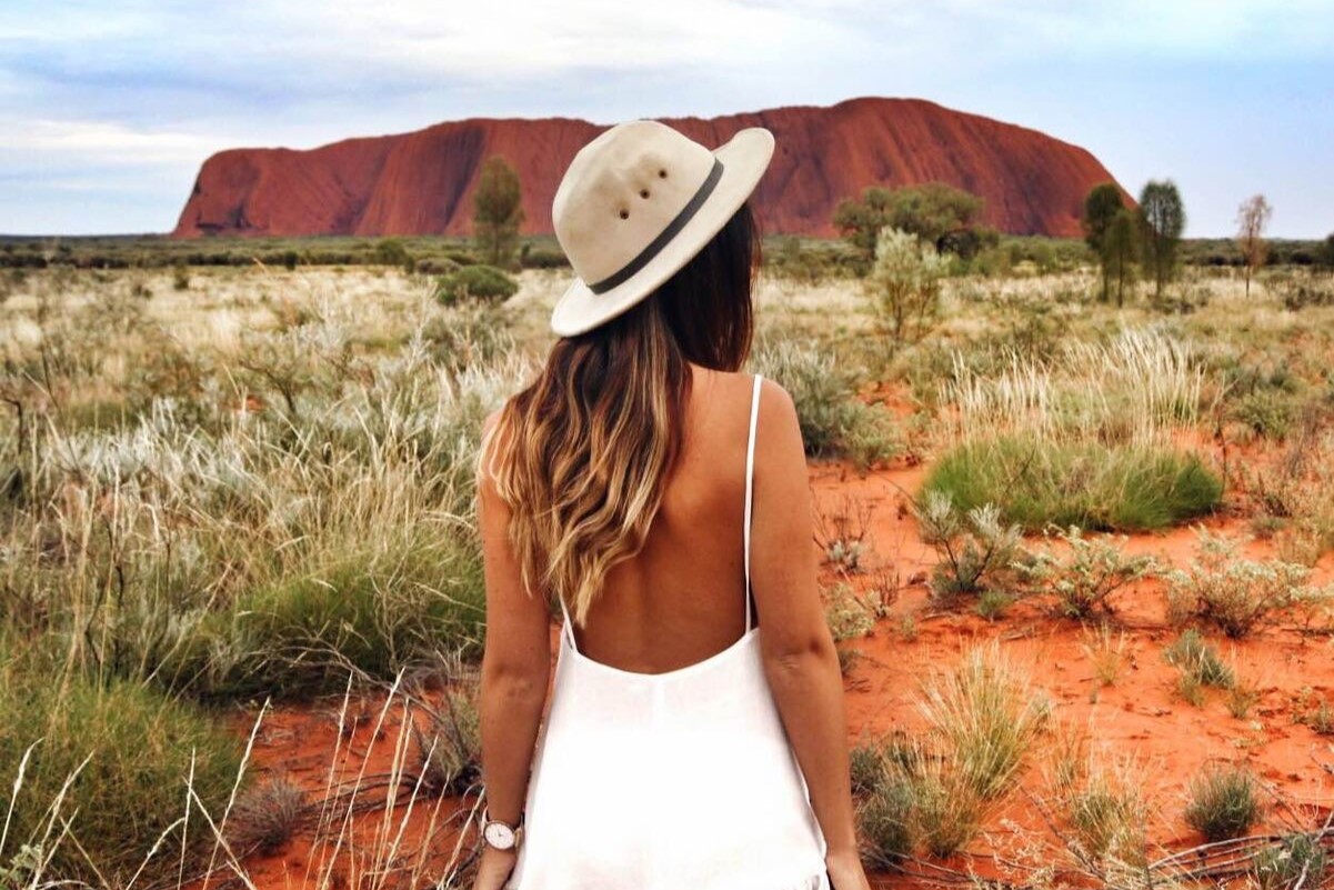 Woman in white dress and hat standing before iconic Uluru during a 3-Day Red Centre Kings Canyon Safari from Alice Springs, Australia.