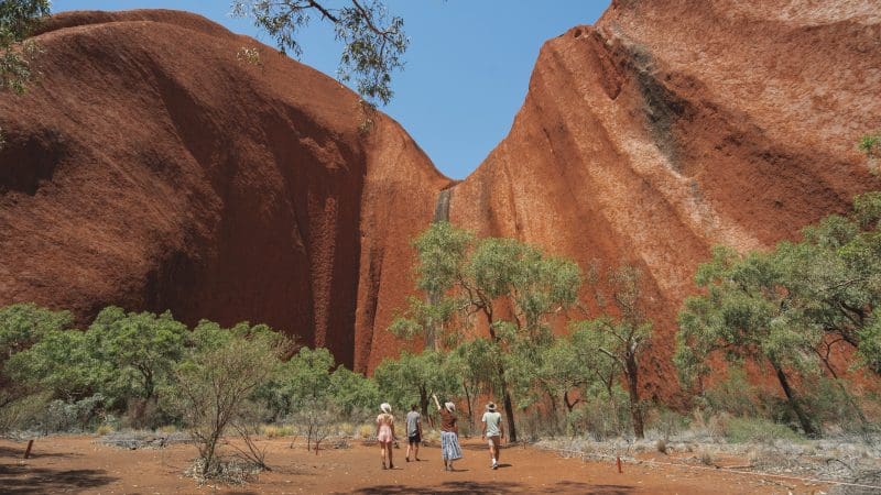 Tour group approaches Uluru on a 4-Day Uluru Kata Tjuta Kings Canyon adventure, blue sky above stunning Australian outback landmarks.