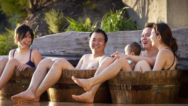 Group of four adults and a baby enjoy relaxing outdoor hot tubs at Peninsula Hot Springs after a one-day spa entry shuttle bus experience.