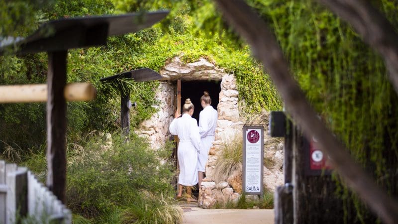 Two guests in white dressing gowns enter a luxurious stone sauna at Peninsula Hot Springs after arriving via the 1 Day Spa Entry Shuttle Bus.