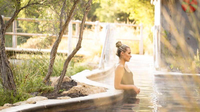 A woman in a stylish black swimming costume unwinds in a natural hot spring after arriving via the Peninsula Hot Springs Spa Entry Shuttle Bus.