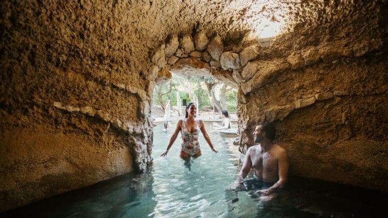 Experience rejuvenation as a woman strolls through a cave’s stone arch and man unwinds nearby; Peninsula Hot Springs shuttle bus outside.