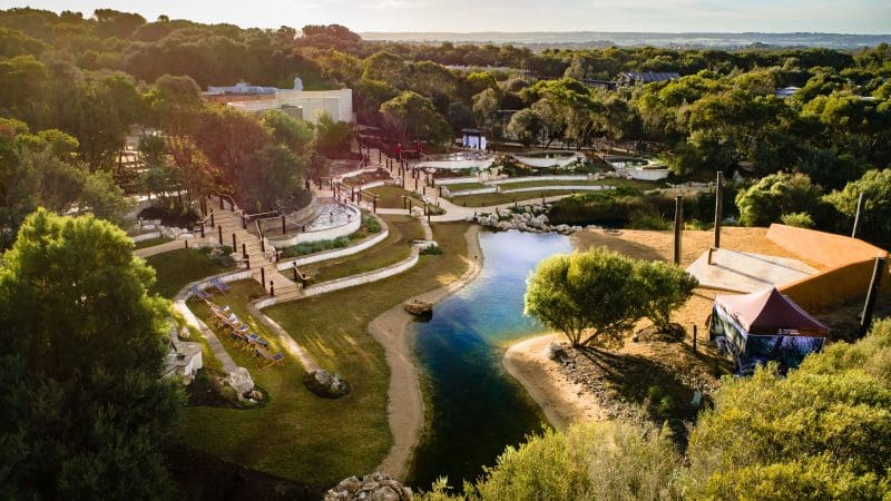 Aerial view of lush park with winding path, serene pond, leafy trees, and visitors—ideal prelude to your Peninsula Hot Springs Spa shuttle.