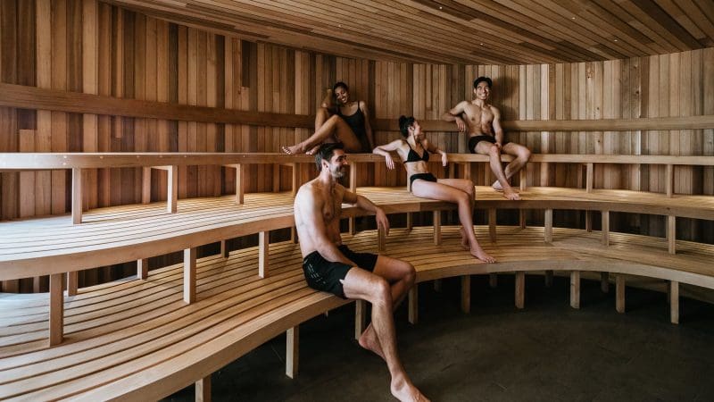 Four guests in swimwear unwind on benches inside a contemporary sauna after enjoying a Peninsula Hot Springs Spa Entry Shuttle Bus experience.