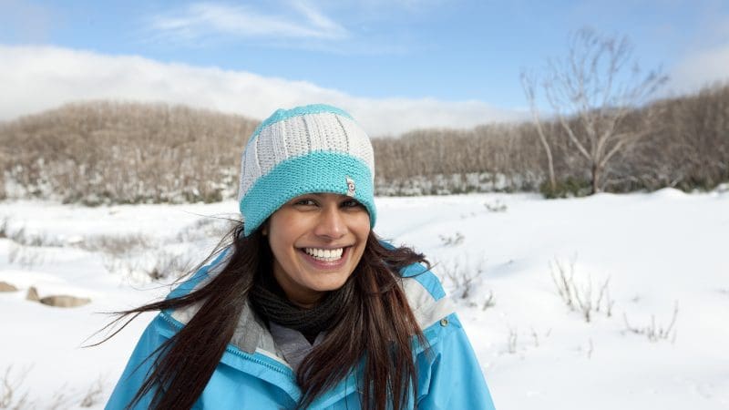 Happy woman in blue jacket enjoying a 1 Day Lake Mountain Snow Waterfalls Tour from Melbourne, with scenic snowy trees behind her.