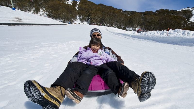 Smiling adult and child in warm winter gear sledging down a snowy hill on a Lake Mountain Snow Tour, near Melbourne, Australia.