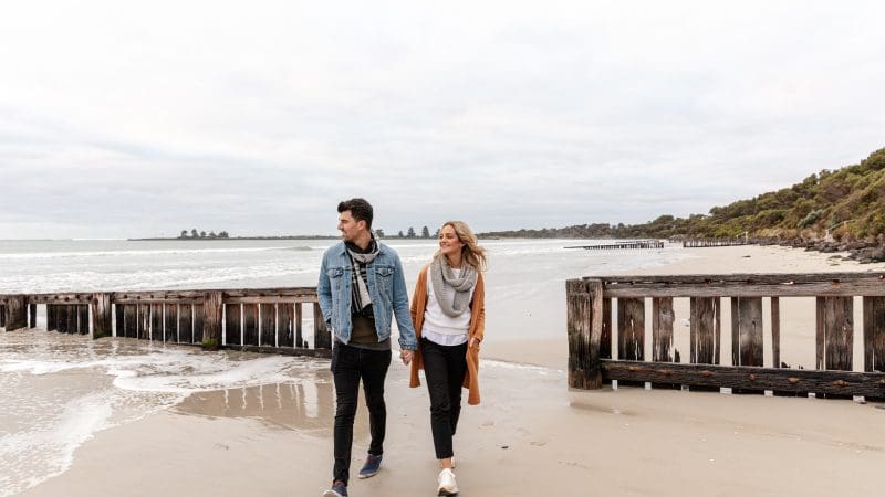 A couple walks along a scenic sandy beach with wooden barriers, capturing the romance of a Great Ocean Road Reverse Eco Tour adventure.