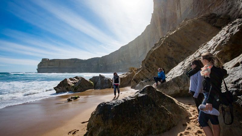 Four travellers unwind on rocky shores beside sandy cliffs and crashing waves on a 1 Day Great Ocean Road Sunset Tour from Melbourne.