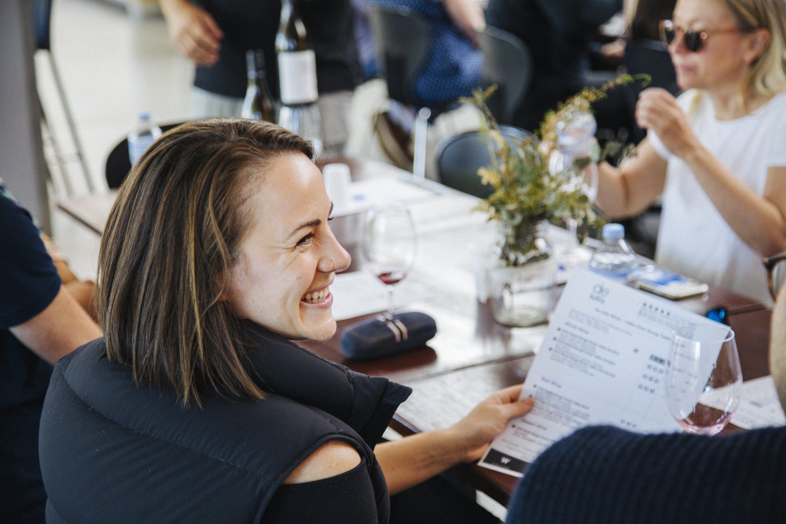 A woman smiles, holding a menu with friends and wine, enjoying a memorable Hunter Valley Signature Wine Tour experience together.