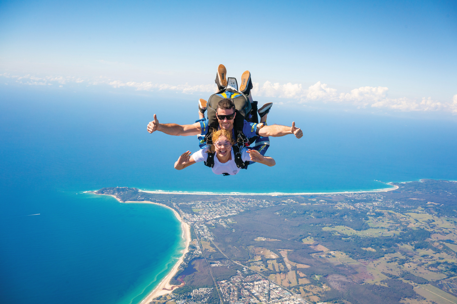 Tandem skydivers above Byron Bay coast, smiling and giving thumbs up during 15,000ft skydive with Brisbane transfer included.