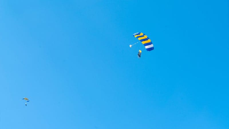 Skydivers with vibrant striped parachutes glide over Wollongong in a tandem skydive from 15,000 feet beneath a clear blue sky.