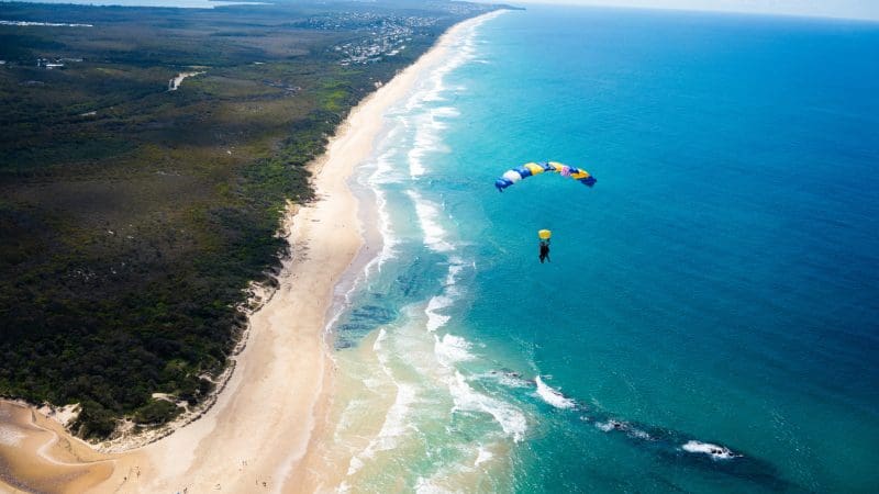 Skydiver with vibrant parachute from Skydive Noosa glides over pristine sandy beach and crystal-clear turquoise sea water below.