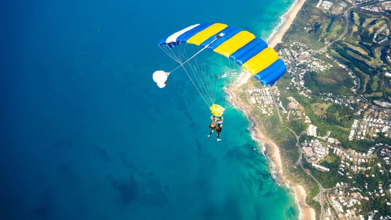 A skydiver leaps with Skydive Noosa from 15,000ft, soaring above turquoise ocean, golden beaches, and the vibrant cityscape below.