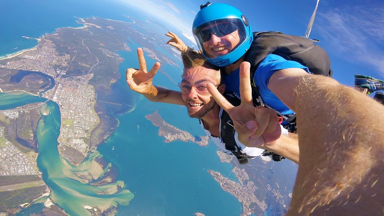 Skydivers tandem jump over Newcastle coast from 15,000 feet, smiling and flashing peace signs in exhilarating skydiving adventure.