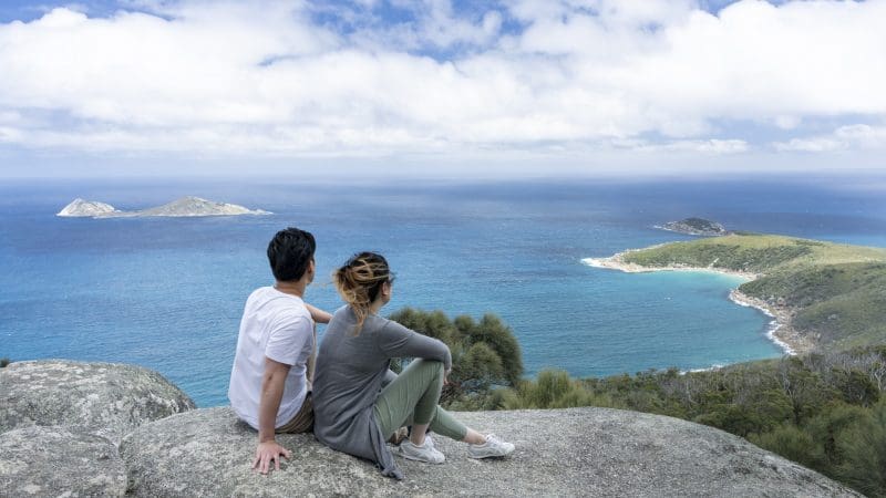 A couple relaxes atop a rocky cliff at Wilsons Promontory Day Tour, overlooking scenic sea views, islands, and lush coastline.