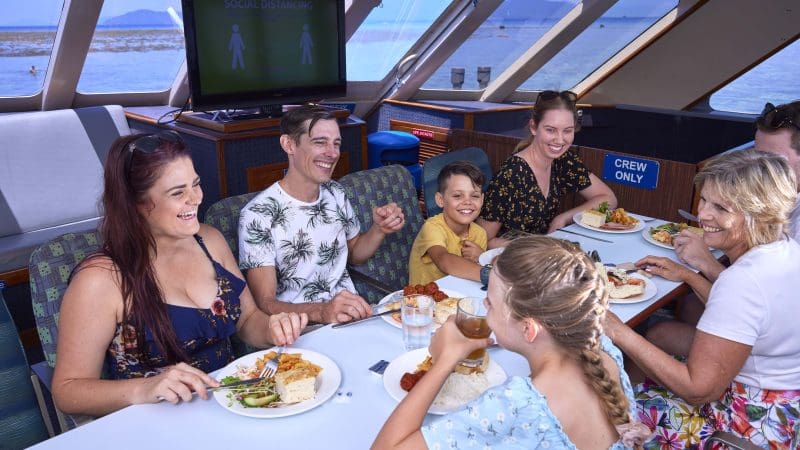 Happy family enjoys a meal and laughter aboard a Full Day Green Island 11AM departure boat, with social distancing sign visible.