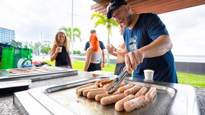 Man barbecuing sausages outdoors at 28 Day Oz East Coast Adventure Intro Travel event, highlighting group travel experience.