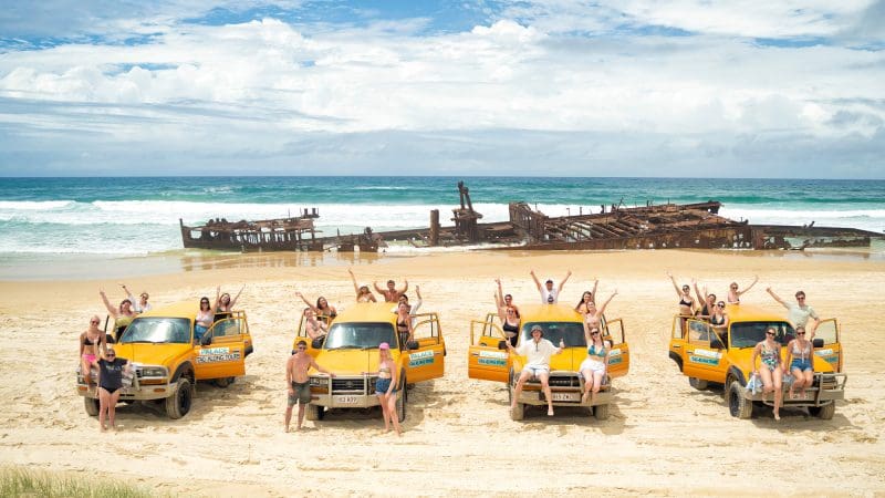 Travellers on the 28 Day Oz East Coast Adventure smile and wave beside vibrant yellow lorries at a famous rusted shipwreck in Australia.