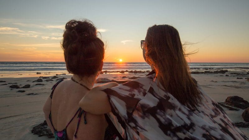 Two women watch a vibrant sunset on a rugged beach, envisioning their unforgettable 10 Day Broome to Darwin 4WD Kimberley Adventure.