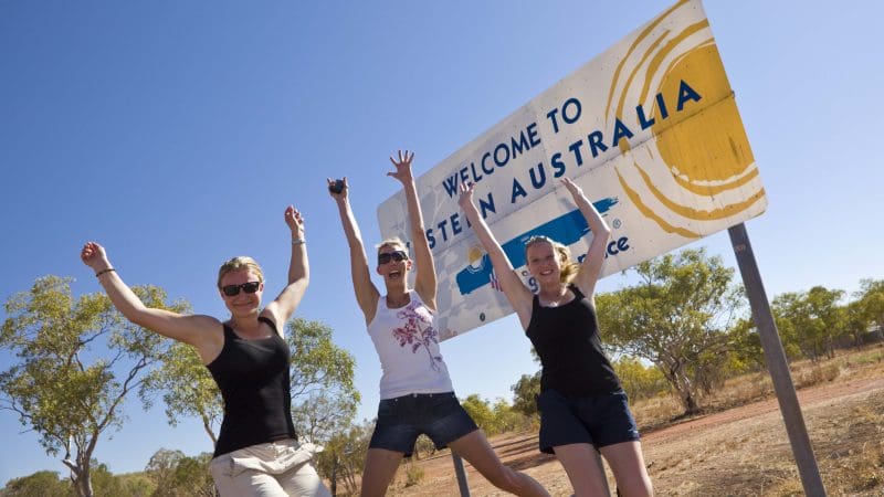 Three happy travellers jump and smile by the iconic Welcome to Western Australia sign on their epic 10 Day Broome to Darwin 4WD Kimberley tour.