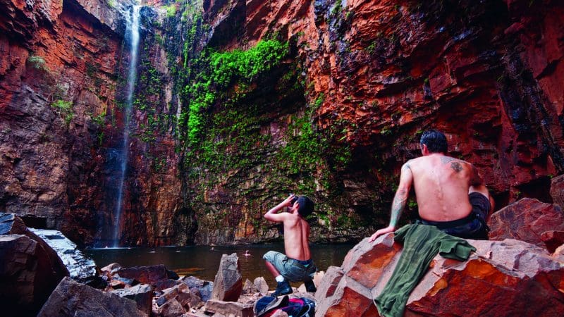 Adventurers rest on rocky terrain beside a cascading waterfall, surrounded by red mossy cliffs during a 10 Day Darwin to Broome 4WD tour.