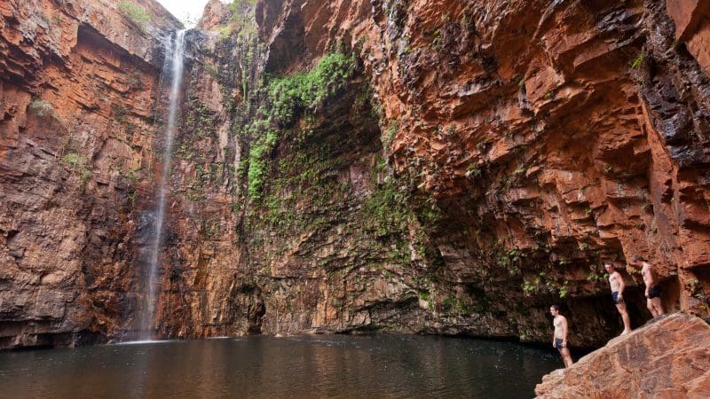 Spectacular waterfall cascades into a rocky pool as Darwin to Broome 4WD Adventure travellers enjoy panoramic views from red cliffs above.