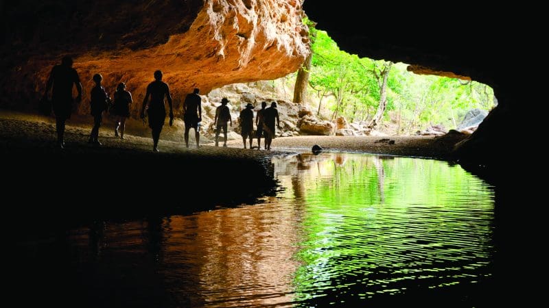 Adventurers silhouetted in a cave by sparkling water on a 10 Day Darwin to Broome 4WD Tour, gazing at lush green trees outside.