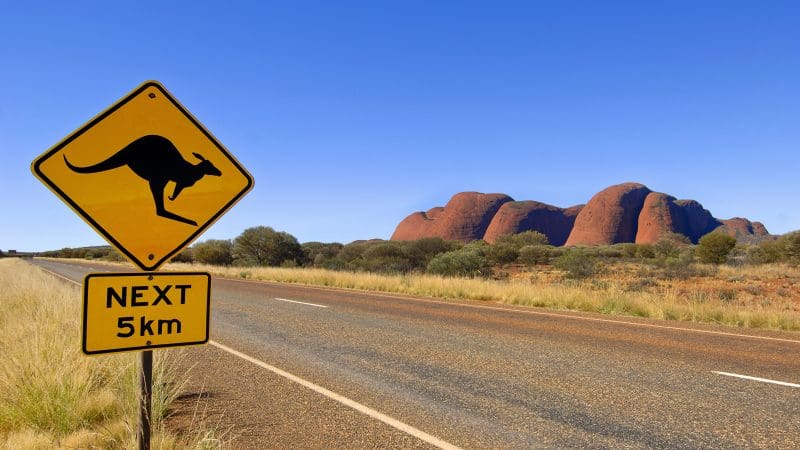 Bright yellow kangaroo road sign with "NEXT 5km" on scenic Uluru tour, Yulara loop; iconic red rock formations in the background.