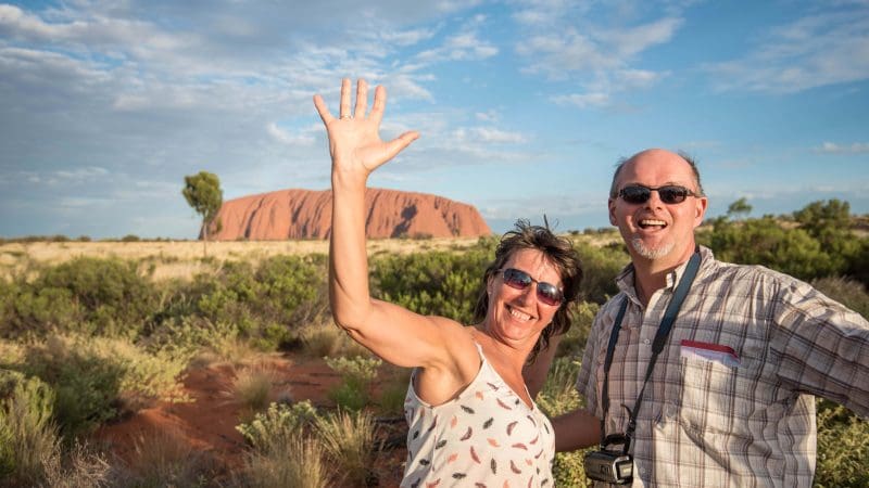Happy couple waving and smiling in front of Uluru on a 3-day Uluru Adventure tour from Alice Springs, enjoying iconic Australian scenery.
