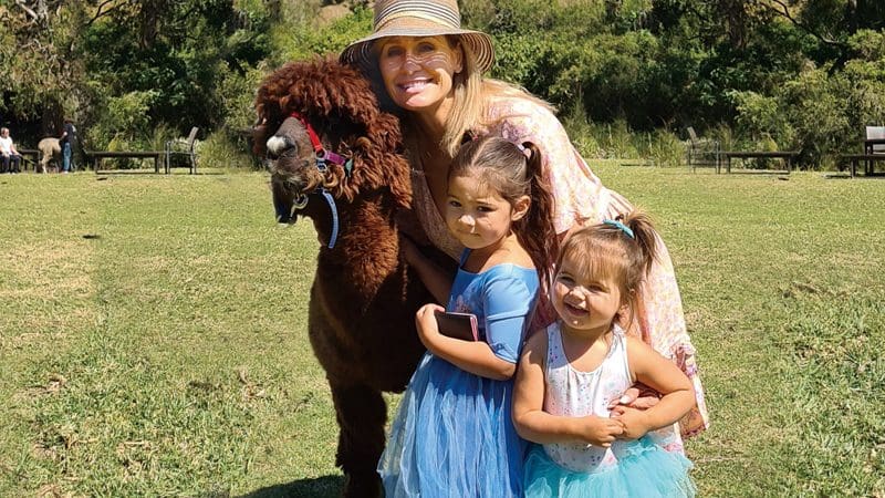 Smiling woman and two girls in blue dresses pose with a brown alpaca at O'Reilly's Lamington National Park near Gold Coast, Australia.