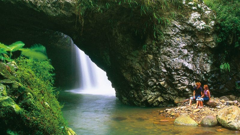 A couple relaxes on rocky terrain beside a stunning cave waterfall, inspired by the Natural Bridge Glow Worm Night Tour Gold Coast.