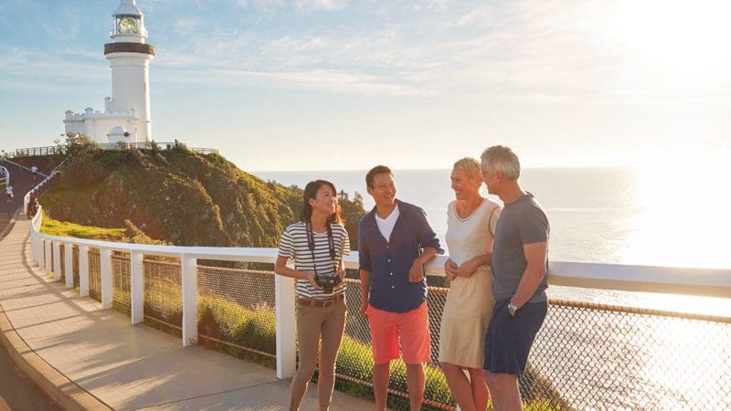 Group of friends relax by Byron Bay lighthouse at sunset, smiling and enjoying breathtaking coastal views of the ocean and landscape.