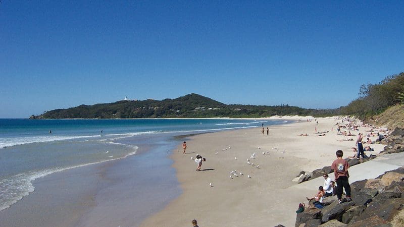 Tourists relaxing on Byron Bay’s pristine sandy beach, enjoying sunshine, gentle waves, and a clear blue sky for the perfect holiday.