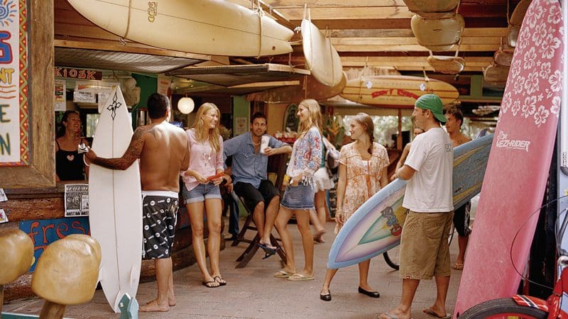 Smiling surfers with colourful boards gather and laugh by a rustic surf shop in Byron Bay, Australia’s top destination for surfing enthusiasts.