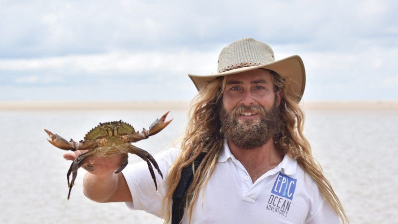 Smiling man in a hat holds a crab by the Noosa Beach shore, wearing a white EPC Ocean Adventures shirt, enjoying coastal adventure.