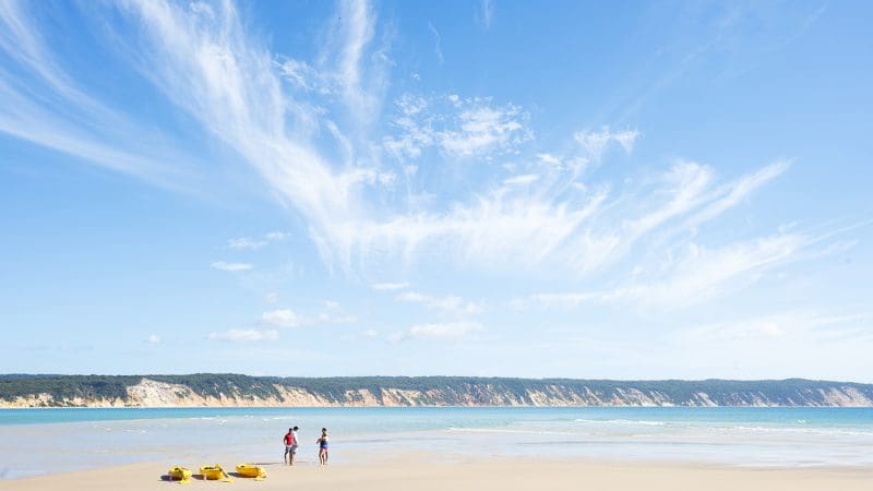 Explore Noosa’s stunning coastline as two adventurers stroll along a sandy beach beside bright yellow kayaks on a Dolphin Sea Kayak 4X4 Tour.