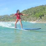 A woman in a vibrant red shirt rides Australia’s Longest Wave by a scenic sandy beach dotted with trees and parked cars in the background.